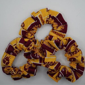 May include: Three scrunchies in a repeating pattern of gold and maroon. The fabric features the word "CHIPPEWAS" and a logo. These hair accessories are a stylish way to keep hair out of the face.