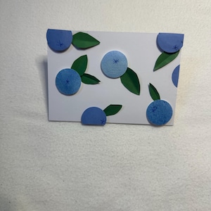 May include: A rectangular white greeting card decorated with several circular blue paper cutouts representing blueberries and pointed green paper cutouts representing leaves. Each blue circle features a small hand-drawn asterisk detail.