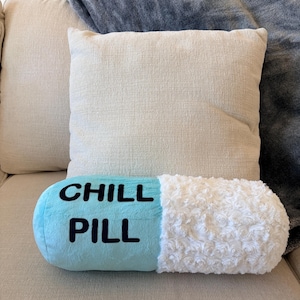 May include: A novelty, pill-shaped cushion with the words "CHILL PILL" in black on a light blue base. One end is white and fluffy. The cushion is on a beige sofa, with two square cushions and a grey blanket in the background.