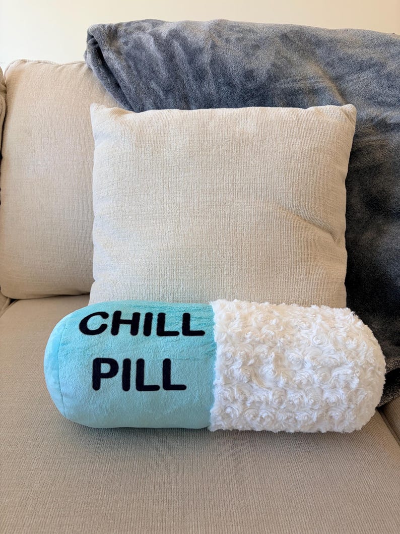 May include: A novelty, pill-shaped cushion with the words "CHILL PILL" in black on a light blue base. One end is white and fluffy. The cushion is on a beige sofa, with two square cushions and a grey blanket in the background.