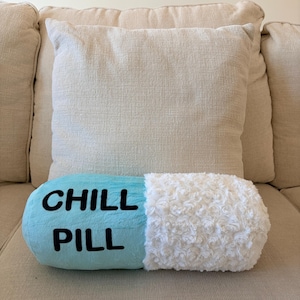 May include: A novelty pillow shaped like a blue and white pill, with the words "CHILL PILL" in black lettering. The pillow is resting on a beige sofa, with a beige cushion in the background.