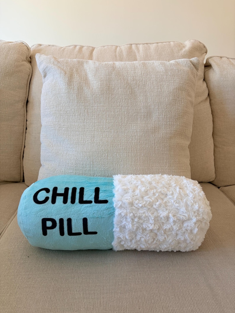 May include: A novelty pillow shaped like a blue and white pill, with the words "CHILL PILL" in black lettering. The pillow is resting on a beige sofa, with a beige cushion in the background.