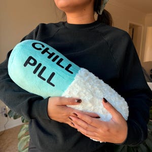 May include: A novelty cushion in the shape of a pill, featuring a light blue top and a fluffy white base. The words "CHILL PILL" are printed in black on the blue section. The cushion is being held by someone.