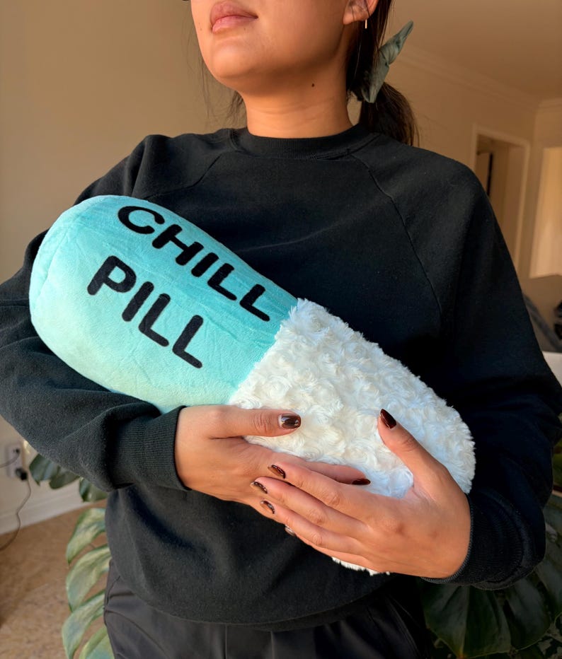 May include: A novelty cushion in the shape of a pill, featuring a light blue top and a fluffy white base. The words "CHILL PILL" are printed in black on the blue section. The cushion is being held by someone.