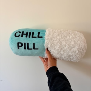 May include: A novelty cushion in the shape of a capsule, with the words "CHILL PILL" in black on a light blue section. One end is white and fluffy. The cushion is held up against a white wall.