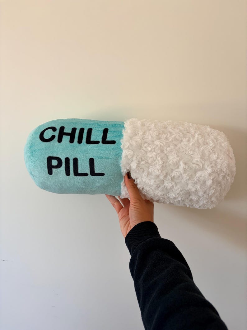 May include: A novelty cushion in the shape of a capsule, with the words "CHILL PILL" in black on a light blue section. One end is white and fluffy. The cushion is held up against a white wall.