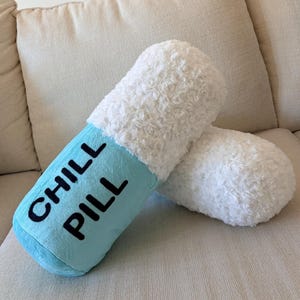 May include: A novelty pillow shaped like a capsule, with a light blue central section and fluffy white ends. The words "CHILL PILL" are printed in black. The pillow is on a beige sofa.