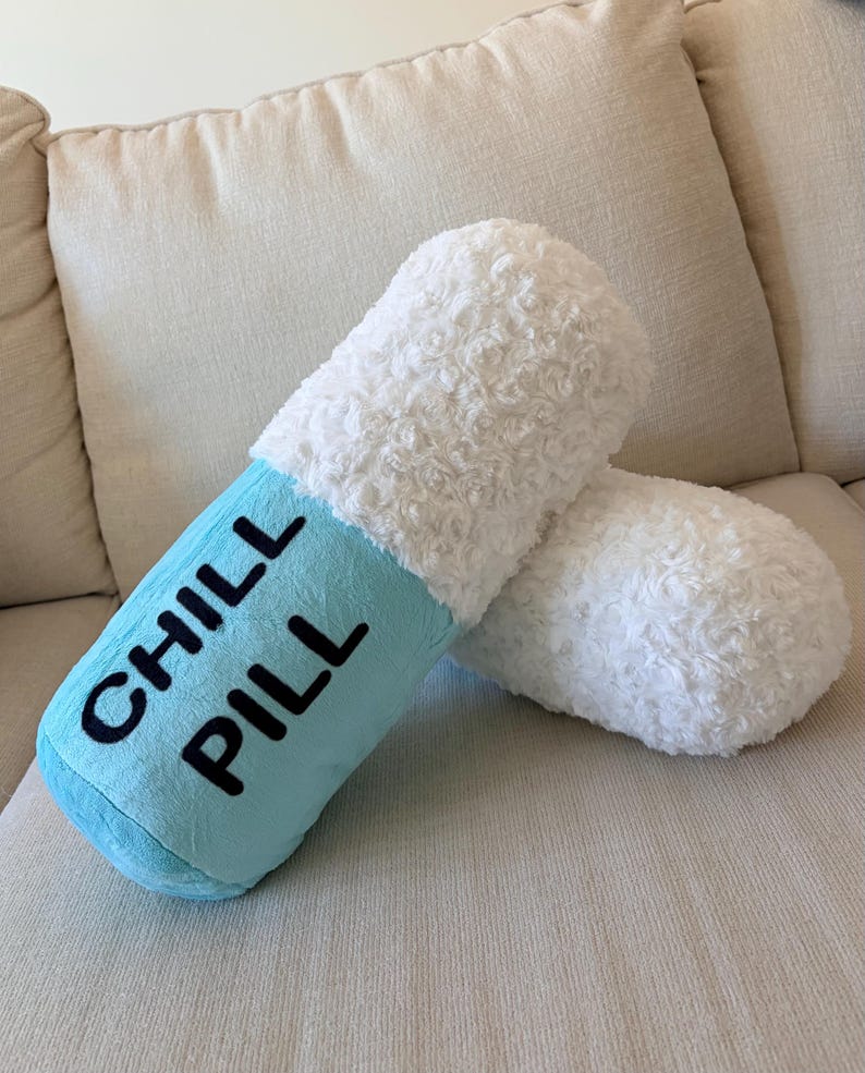 May include: A novelty pillow shaped like a capsule, with a light blue central section and fluffy white ends. The words "CHILL PILL" are printed in black. The pillow is on a beige sofa.