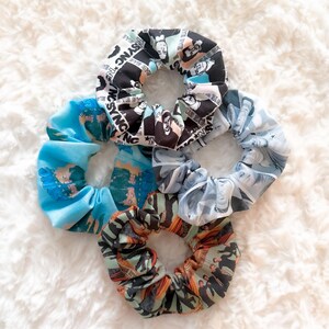 May include: A collection of five fabric scrunchies in various patterns and colors. One scrunchie features a black and white design with text and faces, while others display blue, gray, and orange patterns. These hair accessories are arranged on a white, fluffy surface.