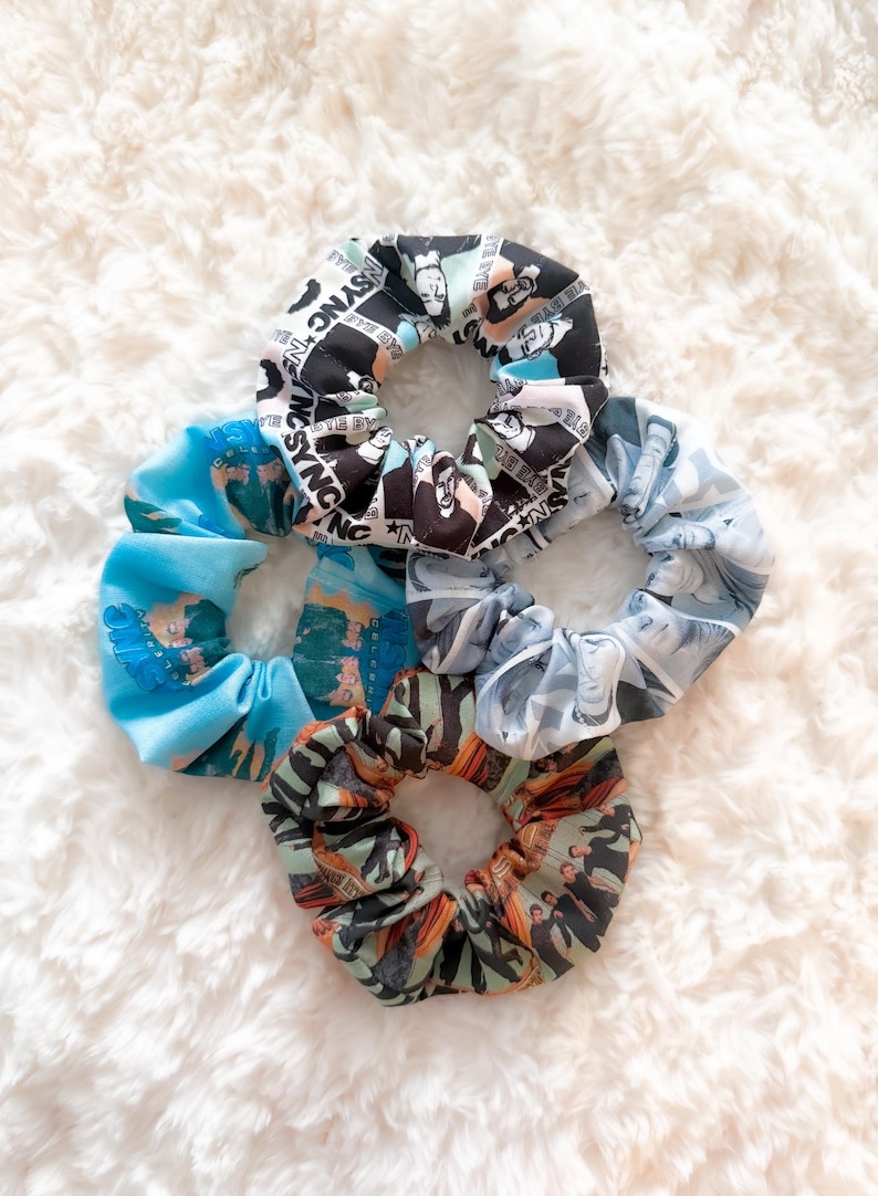 May include: A collection of five fabric scrunchies in various patterns and colors. One scrunchie features a black and white design with text and faces, while others display blue, gray, and orange patterns. These hair accessories are arranged on a white, fluffy surface.