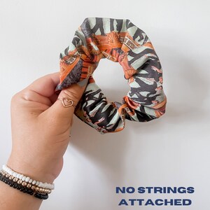 May include: A patterned scrunchie with orange, black, and teal accents. The scrunchie features a repeating design with text. The words "NO STRINGS ATTACHED" are printed in blue on the white background.