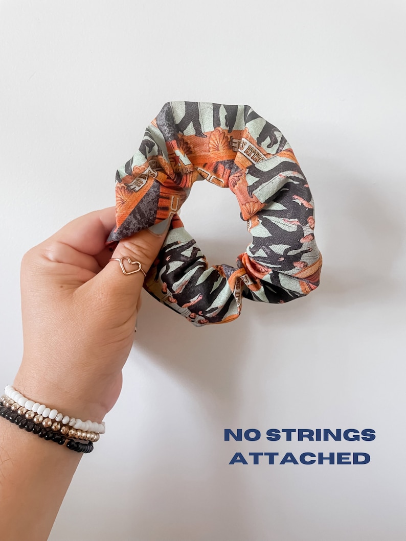 May include: A patterned scrunchie with orange, black, and teal accents. The scrunchie features a repeating design with text. The words "NO STRINGS ATTACHED" are printed in blue on the white background.