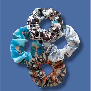 May include: A collection of five fabric scrunchies in various patterns and colors. One scrunchie features a black and white design with the text "NSYNC". Other scrunchies have blue, gray, and orange patterns. The background is a solid blue.