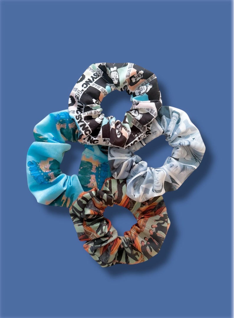 May include: A collection of five fabric scrunchies in various patterns and colors. One scrunchie features a black and white design with the text "NSYNC". Other scrunchies have blue, gray, and orange patterns. The background is a solid blue.