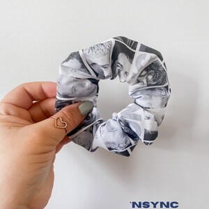 May include: A black and white scrunchie featuring a collage of faces. The scrunchie is held by a hand with a gold heart ring. The word "NSYNC" is printed in blue at the bottom.