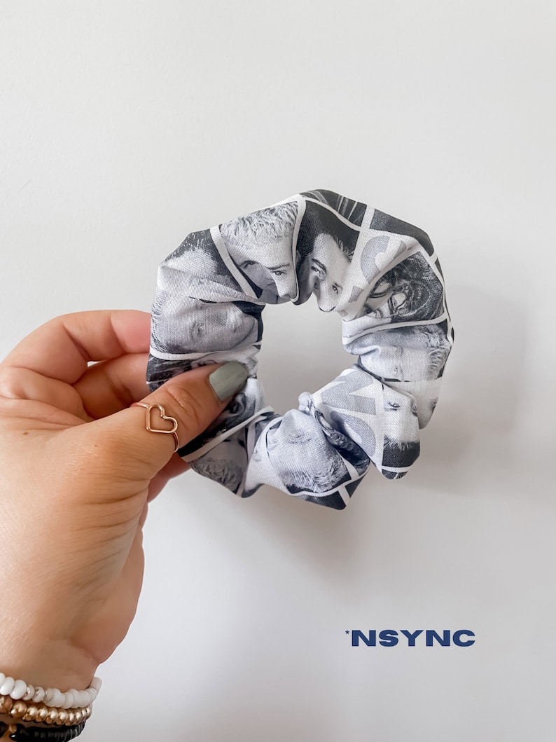 May include: A black and white scrunchie featuring a collage of faces. The scrunchie is held by a hand with a gold heart ring. The word "NSYNC" is printed in blue at the bottom.