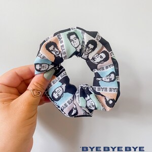 May include: A scrunchie with a repeating pattern of black and white portraits on a pastel background. The text "BYE BYE BYE" is printed on the scrunchie. The scrunchie is held by a hand with a gold heart ring.