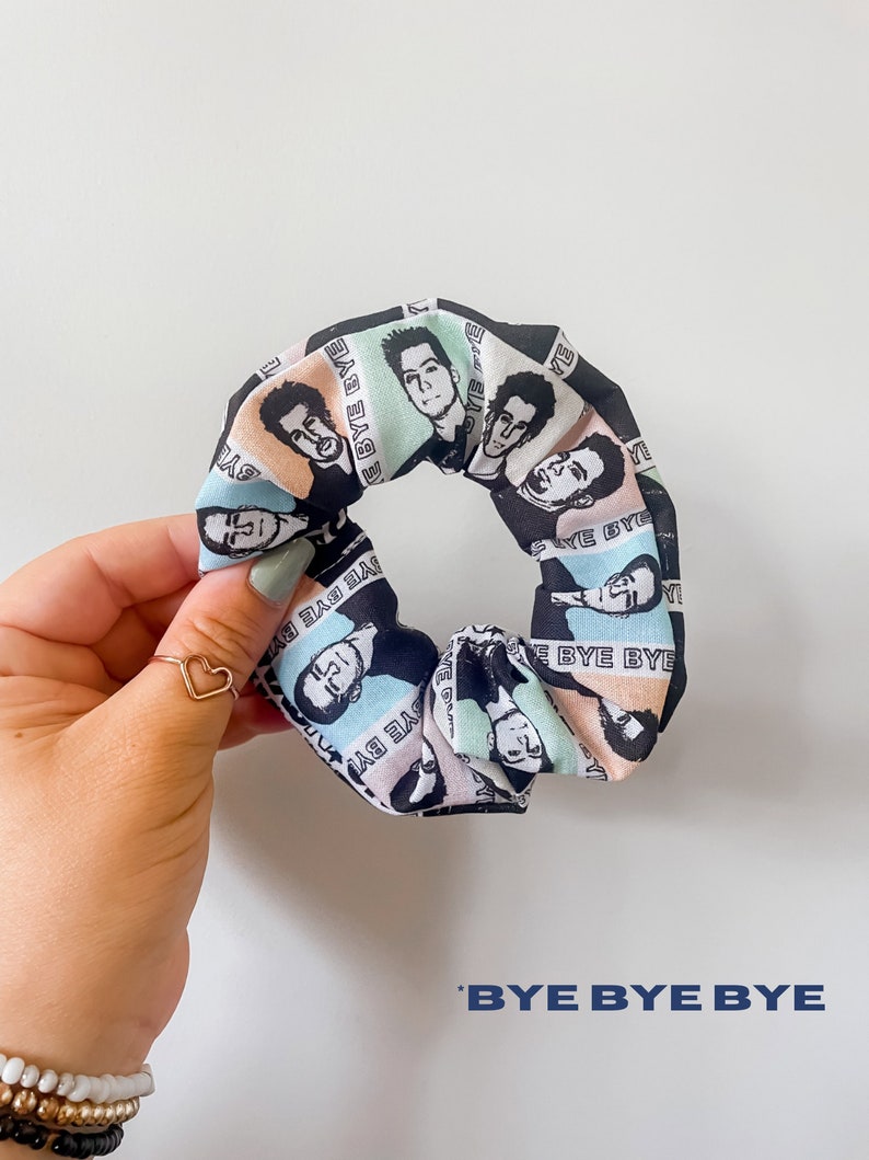 May include: A scrunchie with a repeating pattern of black and white portraits on a pastel background. The text "BYE BYE BYE" is printed on the scrunchie. The scrunchie is held by a hand with a gold heart ring.