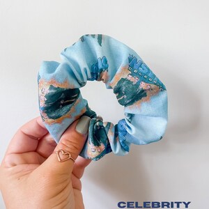 May include: A light blue scrunchie with a repeating pattern featuring a group of people and the word "CELEBRITY". The scrunchie is made of fabric and is a hair accessory.