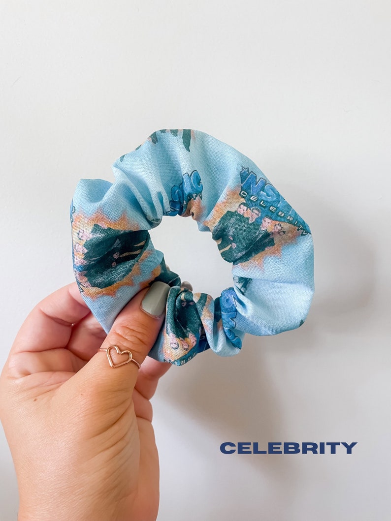 May include: A light blue scrunchie with a repeating pattern featuring a group of people and the word "CELEBRITY". The scrunchie is made of fabric and is a hair accessory.