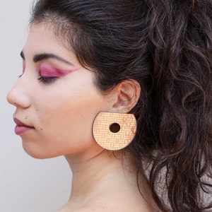 Peut inclure: Une femme aux cheveux bruns longs et bouclés portant une grande boucle d'oreille ronde en bois avec un motif tissé et un trou au centre.