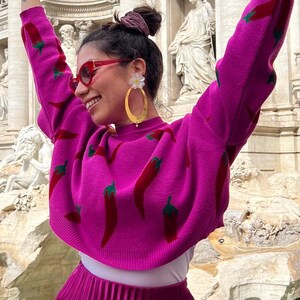 Op de afbeelding: Een magenta trui met een patroon van rode chilipepers. De persoon draagt een rode zonnebril en gouden oorringen. De outfit omvat een magenta geplooide rok.