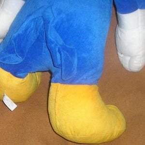 May include: A plush toy featuring a bright blue body, large white gloves, and oversized yellow feet. The character has a blue tail extending from the back. A small white fabric tag is attached to the side of one of the yellow plush feet.