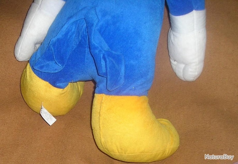 May include: A plush toy featuring a bright blue body, large white gloves, and oversized yellow feet. The character has a blue tail extending from the back. A small white fabric tag is attached to the side of one of the yellow plush feet.