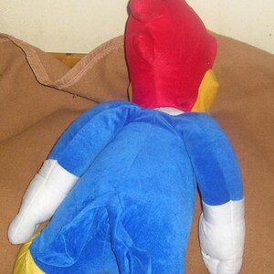 May include: A plush toy of a cartoon woodpecker character featuring a bright red head, a blue body, white gloves, and yellow feet. The character is shown from the back, displaying its blue tail feathers and the distinct red crest on its head.