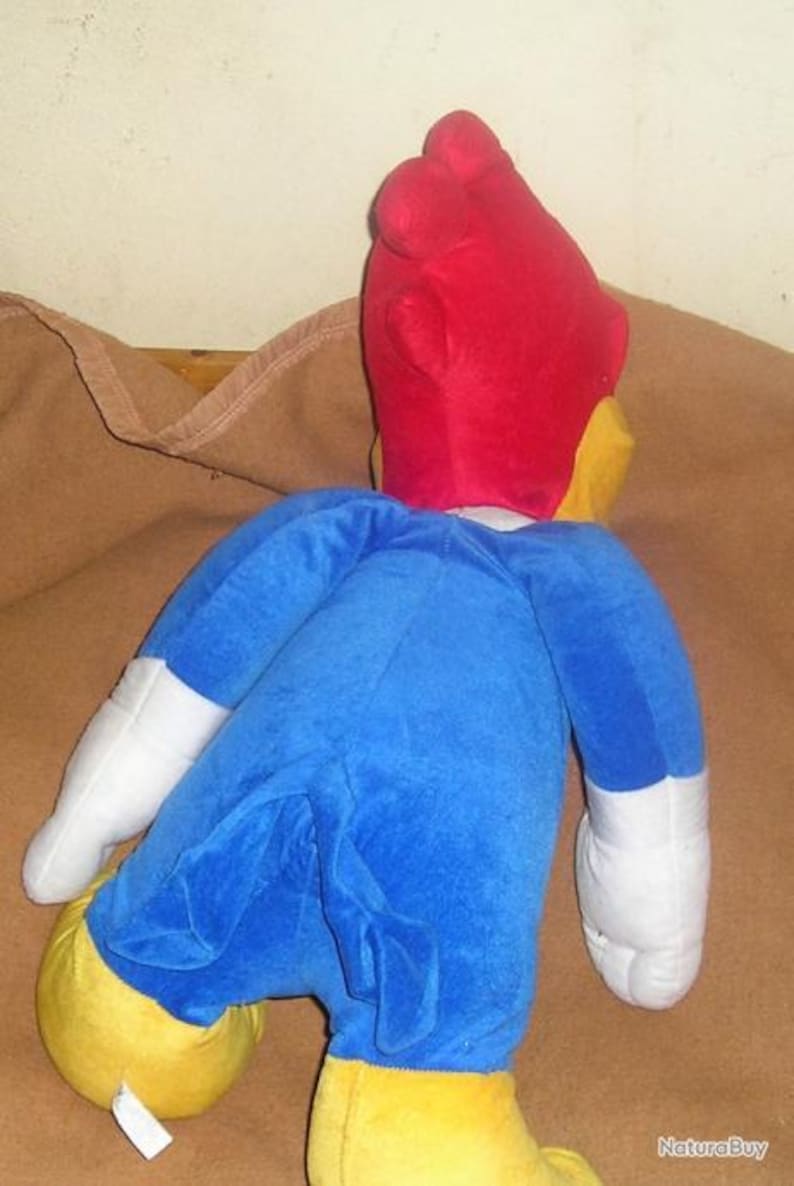May include: A plush toy of a cartoon woodpecker character featuring a bright red head, a blue body, white gloves, and yellow feet. The character is shown from the back, displaying its blue tail feathers and the distinct red crest on its head.