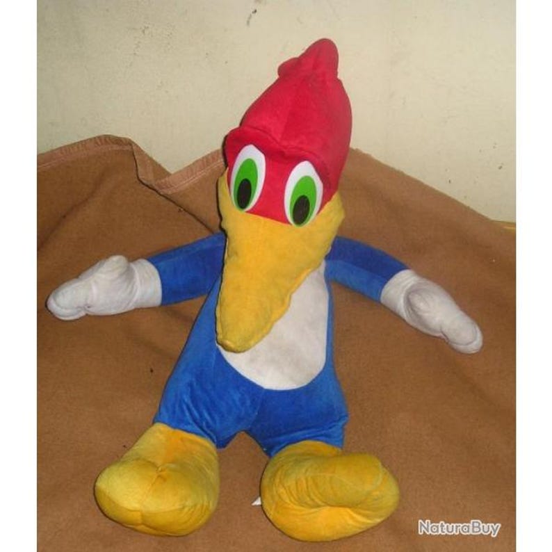 May include: A plush toy of a woodpecker character featuring a bright red crest, a long yellow beak, and large green eyes. The body is blue with a white chest, white gloves on the hands, and yellow feet. The character has a cartoon-like appearance.