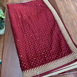 May include: A burgundy fabric with a geometric pattern and gold embellishments. The fabric is folded on a wooden surface. A gold embroidered border runs along the edge of the fabric.