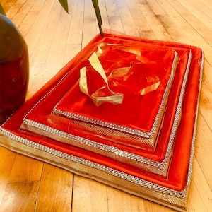 Op de afbeelding: Een stapel van drie rode stoffen geschenkdozen met gouden randen en een gouden lint. De dozen zijn genest, waarbij elke doos iets kleiner is dan de vorige. De dozen staan op een houten oppervlak.