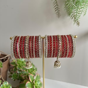 May include: Two sets of red bangles with gold accents and dangling bell charms. The bangles are displayed on a gold stand, showcasing their intricate design. The bangles are adorned with white beads and sparkling embellishments, adding to their elegance.