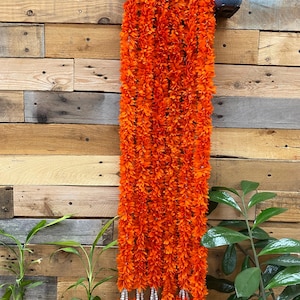 Puede incluir: Una larga y vibrante guirnalda floral naranja con detalles de cuentas blancas cuelga de una rústica pared de madera. La guirnalda está densamente llena de pequeños elementos en forma de flor, creando una apariencia completa y texturizada. La parte inferior de la guirnalda presenta pequeños racimos florales.