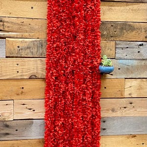 May include: A long, vibrant red floral garland, likely artificial, hangs against a rustic wooden wall. The garland is densely packed with small, red flower-like elements. White accents are visible at the bottom. A small succulent in a blue pot is mounted on the wall.