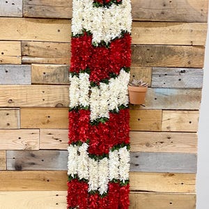 May include: A long, vertical floral garland with alternating sections of red and white flowers, hanging against a rustic wooden wall. A small potted plant sits on the wall. The garland is a decorative item.