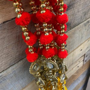 Può includere: Una ghirlanda decorativa con pompon rossi, perline dorate e pendenti dorati ornati con dettagli a specchio. La ghirlanda ha un nappa gialla in basso. Lo sfondo è una superficie di legno.