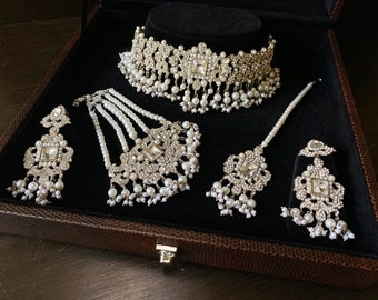 May include: A silver and white bridal jewelry set with a choker necklace, a long necklace, a maang tikka, and earrings. The jewelry is decorated with pearls and rhinestones.