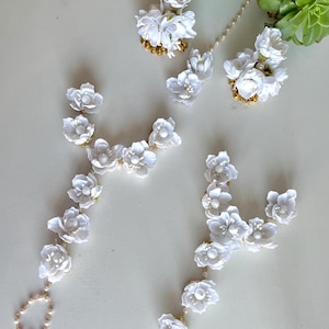 Op de afbeelding: Witte bloemen sieradenset met delicate bloemmotieven. De set bevat een hoofdband, oorbellen en twee handkettingen. De bloemelementen zijn geaccentueerd met kleine gouden kralen en parelachtige details.