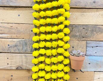 May include: A vertical strand of bright yellow marigold flowers hangs against a rustic wooden background. The flowers are densely packed, creating a vibrant, eye-catching display. A small terracotta pot with a succulent plant is mounted on the wooden surface.