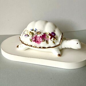 May include: A white ceramic turtle ornament with a floral pattern on its shell. The shell displays pink, purple, and blue flowers with green foliage. The shell's edges are gold-trimmed. The turtle sits on a white, oval-shaped base.