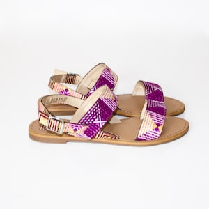 Peut inclure: Une paire de sandales plates en cuir, de couleur violette et marron, avec un motif géométrique. Les sandales ont une boucle de fermeture.