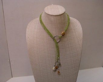 May include: A green beaded necklace with a silver-tone ring and natural stone accents. The necklace is displayed on a mannequin. The necklace has a Y-shaped design with dangling stone pendants.