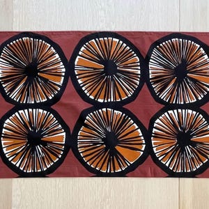 Op de afbeelding: Een rechthoekige tafelloper met een herhalend patroon van cirkelvormige motieven. Elk motief bestaat uit een donkerbruin centrum met uitstralende zwarte en witte lijnen, op een effen terracotta-kleurige achtergrondstof.