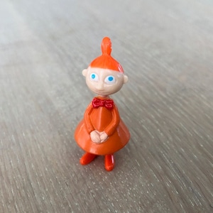 May include: Orange plastic figurine of a girl with red hair, wearing a red bow and an orange dress. The figurine has blue eyes and is standing on a wooden surface.