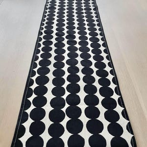 Puede incluir: Un camino de mesa en blanco y negro con un patrón repetitivo de grandes círculos negros sobre un fondo blanco. El camino de mesa está colocado sobre una mesa de madera de color claro. El diseño del camino de mesa añade un toque moderno al comedor.