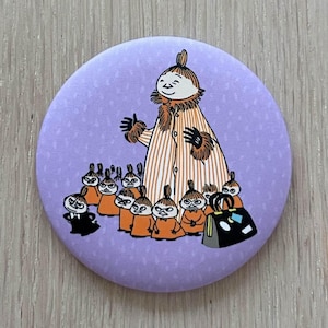 May include: A round, lavender button featuring a cartoon illustration. The central figure, in a striped coat, is surrounded by smaller characters in orange outfits. A black bag with white accents is also present.