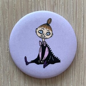 May include: A round, lavender button featuring a cartoon illustration of a character with orange hair in a bun, blue eyes, and a black dress with a pink lining. The character is seated with legs crossed. The button is on a wooden surface.