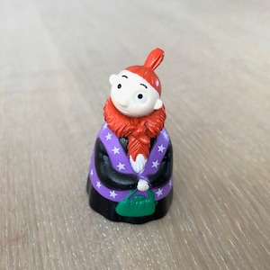 May include: A small figurine of a gnome-like character. The figure has a white face, red hair and beard, and wears a black robe with purple sleeves and white stars. It holds a green purse.